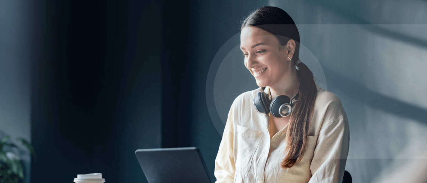 Woman smiling while sitting at her desk, she is wearing headphones around her neck. 